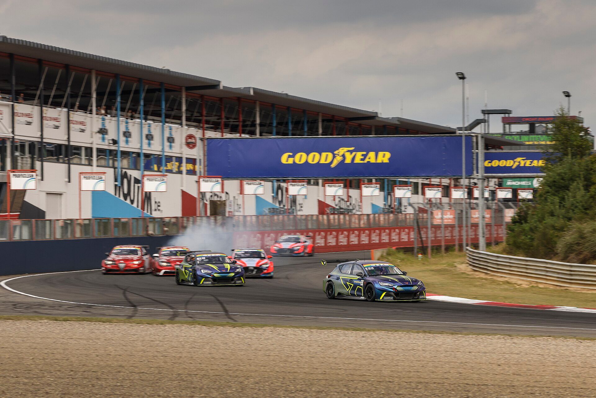 Racing cars in a corner at Circuit Zolder, with the pit building, a grandstand and a banner over the track behind