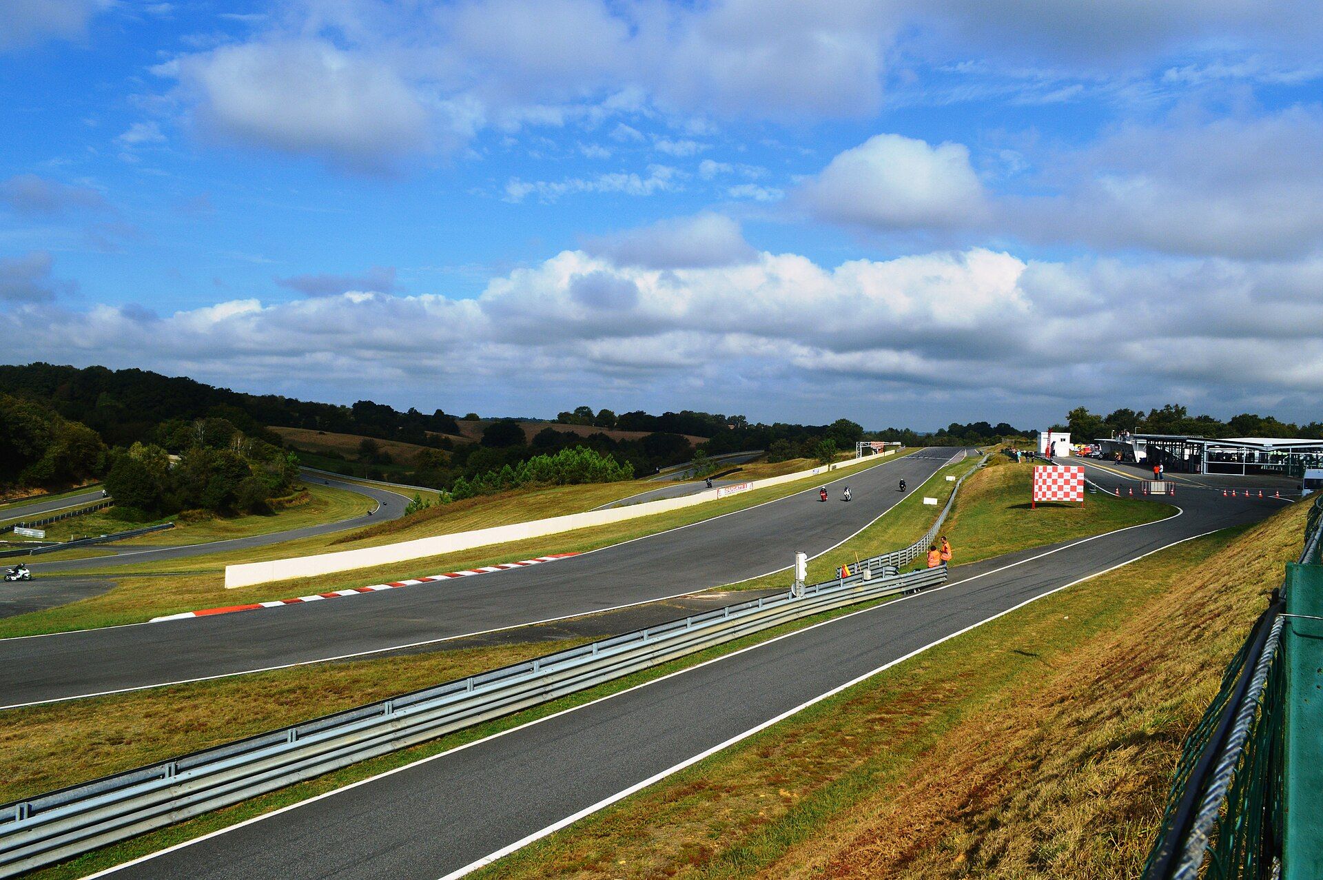 Le circuit de Pau-Arnos vu depuis un talus : ligne droite avec des motos, voie des stands derrière la glissière et bâtiments des stands à droite