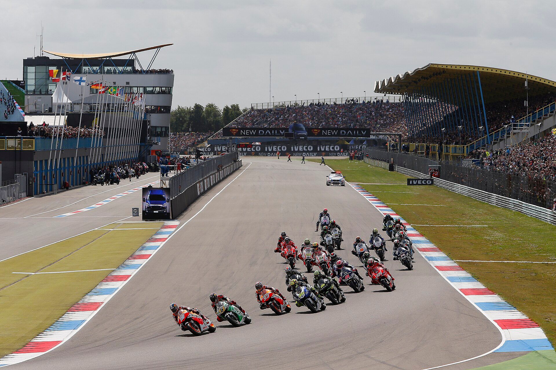 Motorcycles on the main straight of TT Circuit Assen, with a building and tower on the left and a covered grandstand on the right