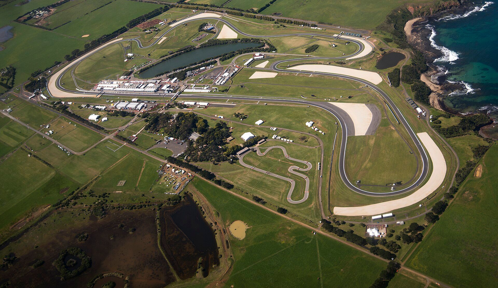 Phillip Island Grand Prix Circuit from the air: the loop among pastures on the ocean shore, with a lake and the pit buildings in the middle