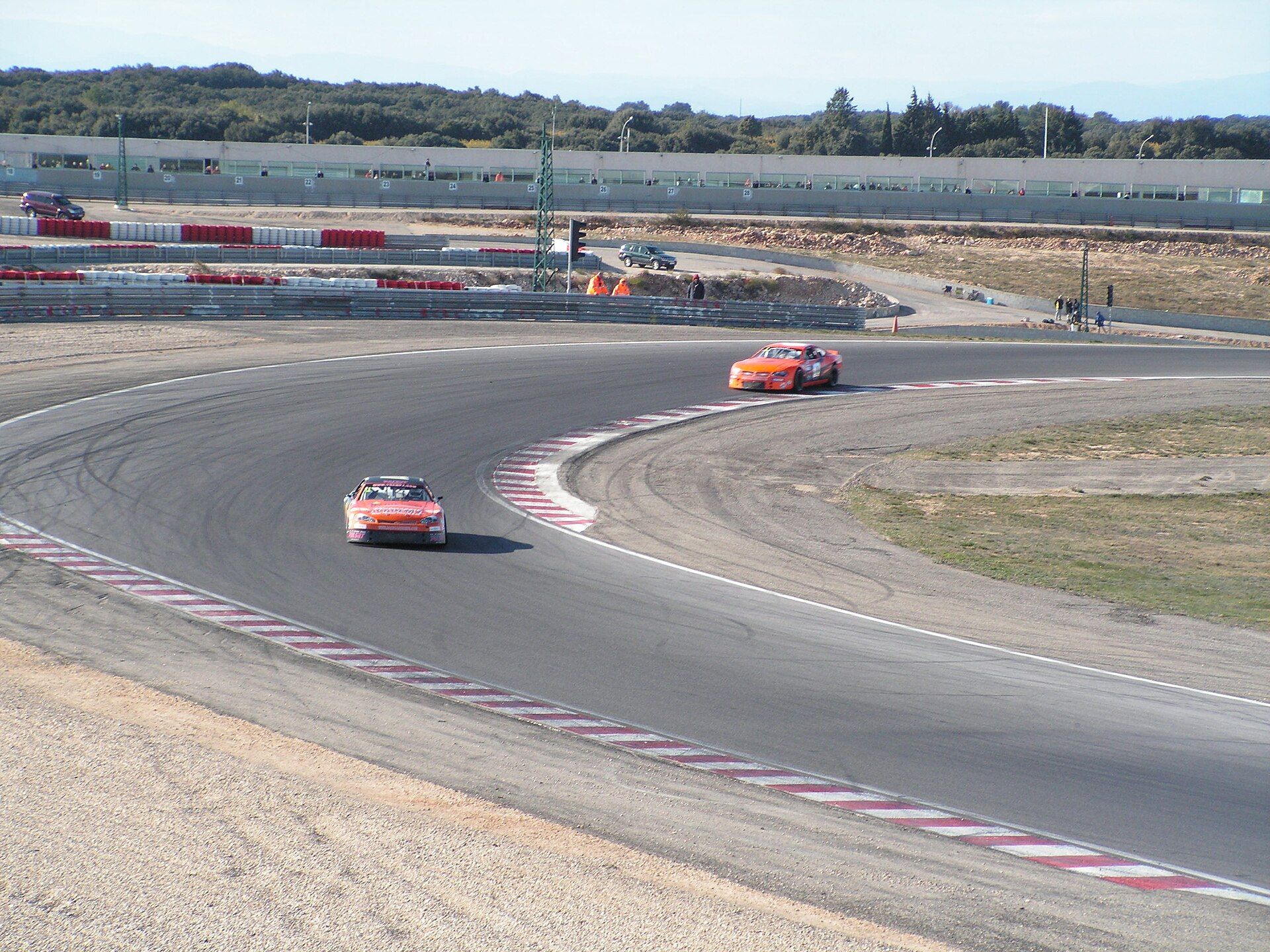 Two racing cars in a left-hand bend of the Lédenon circuit, with barriers and the venue's buildings behind